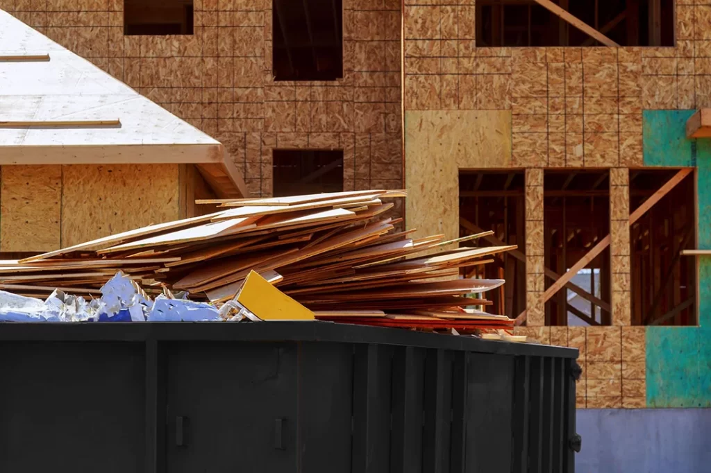 Black roll off dumpster beside stacked lumber at a home construction site
